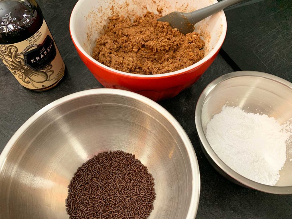 bowl of rum ball mixture beside a bowl of chocolate sprinkles and a bowl of icing sugar