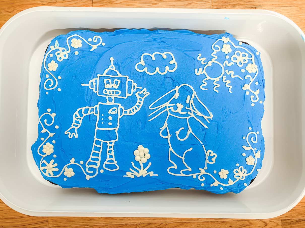 9x13 cake covered with blue frosting, and decorations piped on that look like a robot, a bunny, and some flowers around the edge