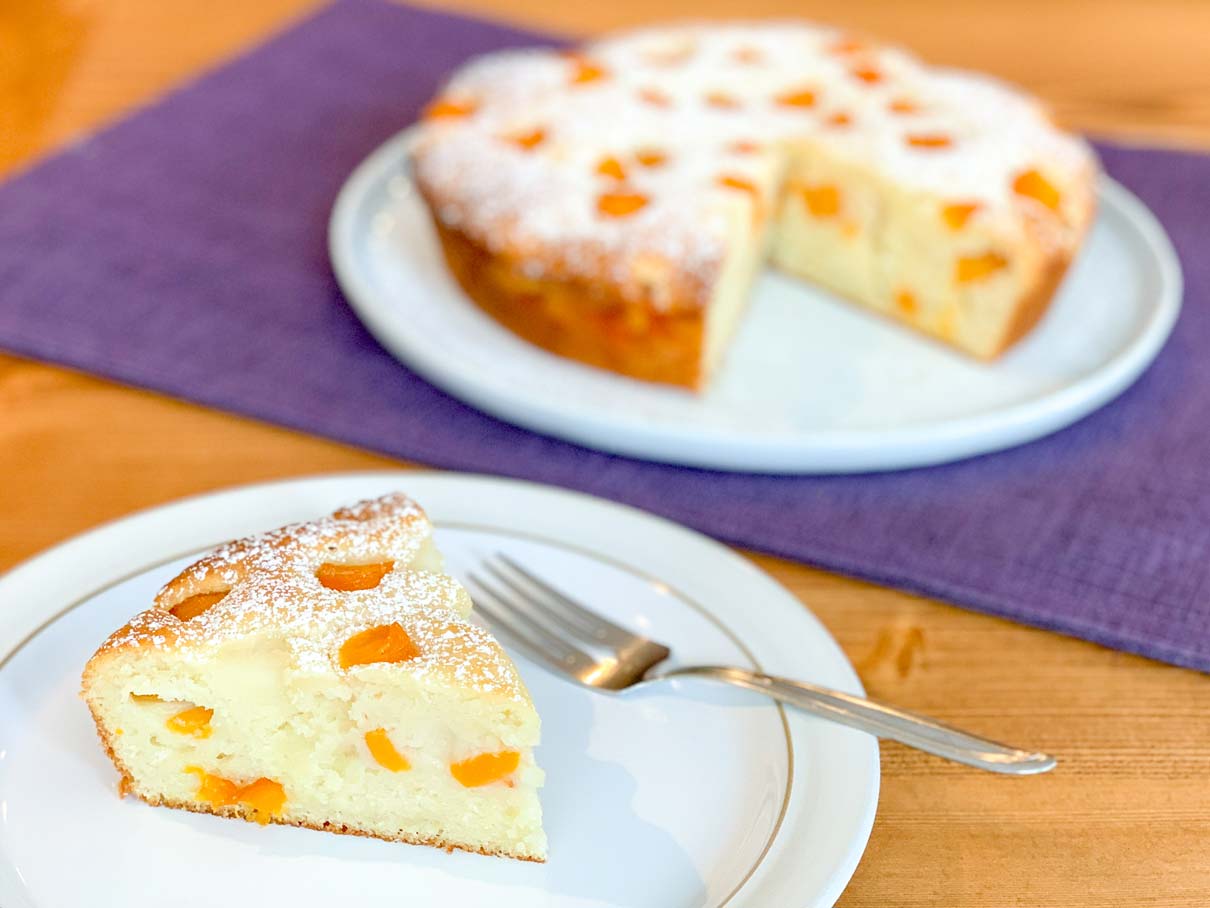 a slice of peach ricotta cake on a plate with the rest of the cake in the background