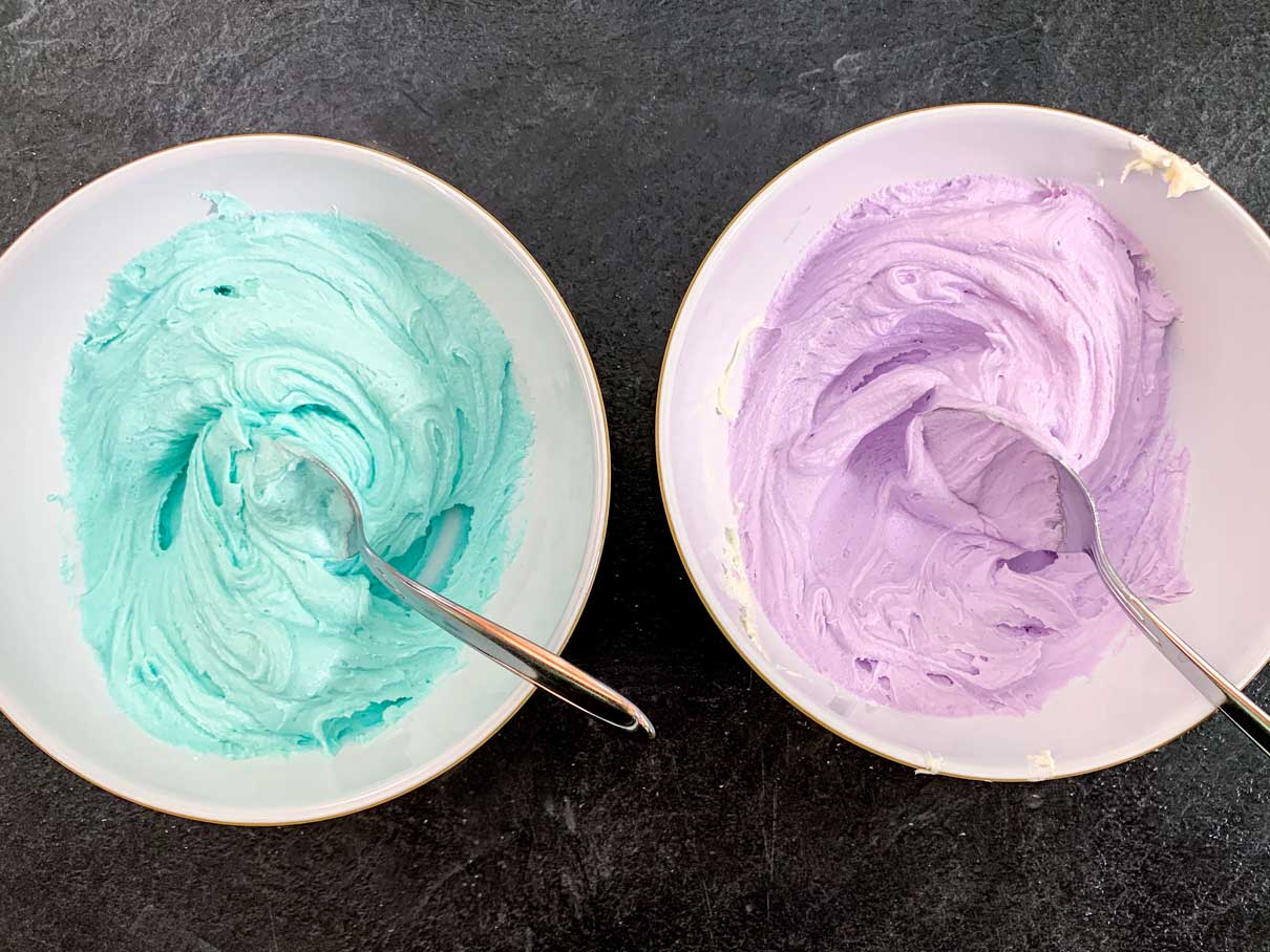 Two bowls of buttercream frosting, one tinted purple, the other tinted blue