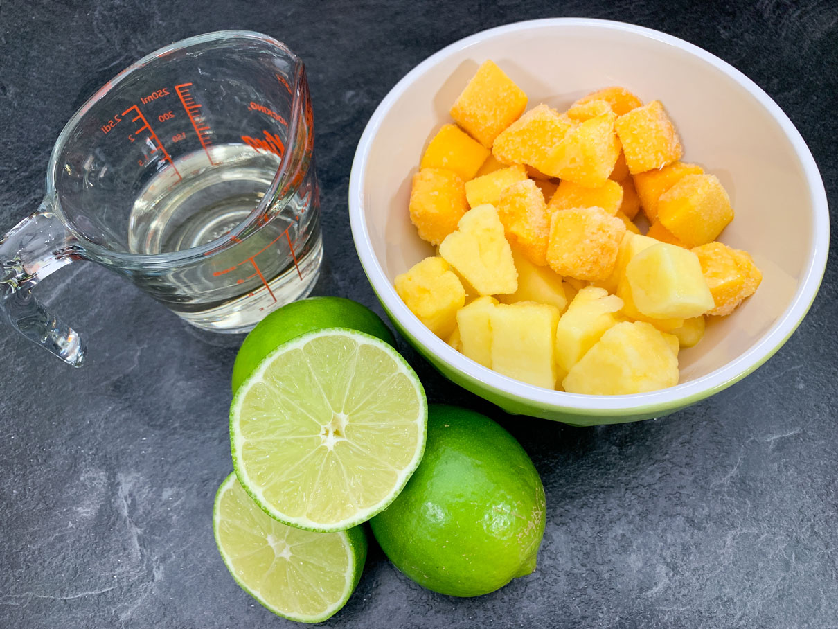 Frozen mango and pineapple in a bowl, next to some limes and tequila.