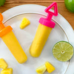 two frozen popsicles on a plate next to limes and pineapple pieces