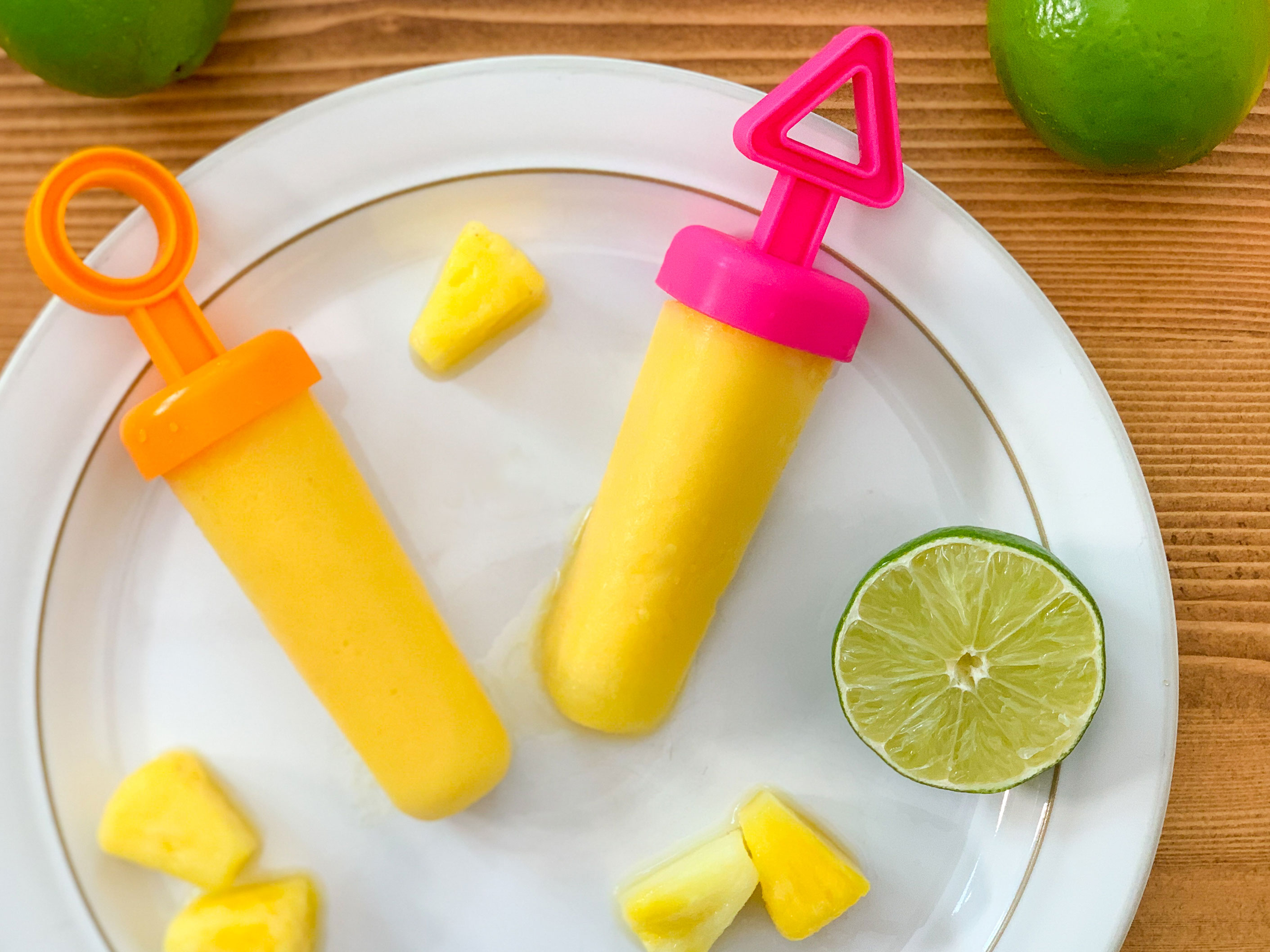 two frozen popsicles on a plate next to limes and pineapple pieces
