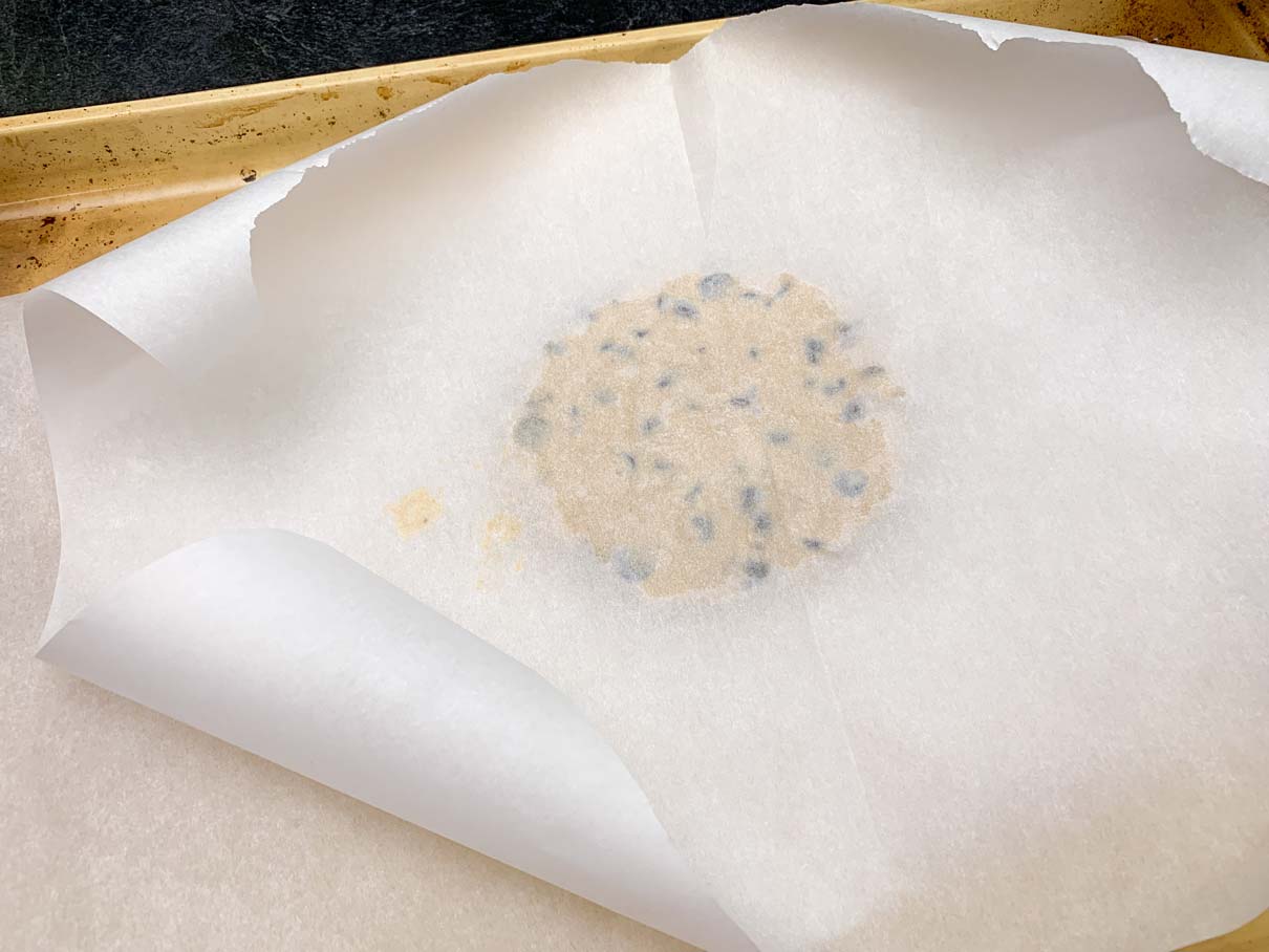 a ball of cookie dough on a baking sheet, with a piece of parchment paper on top, partially flattened