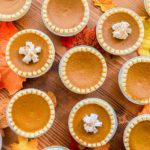 overhead shot of mini pumpkin pies, topped with whipped cream and cinnamon, on wood table top, surrounded by fall leaves