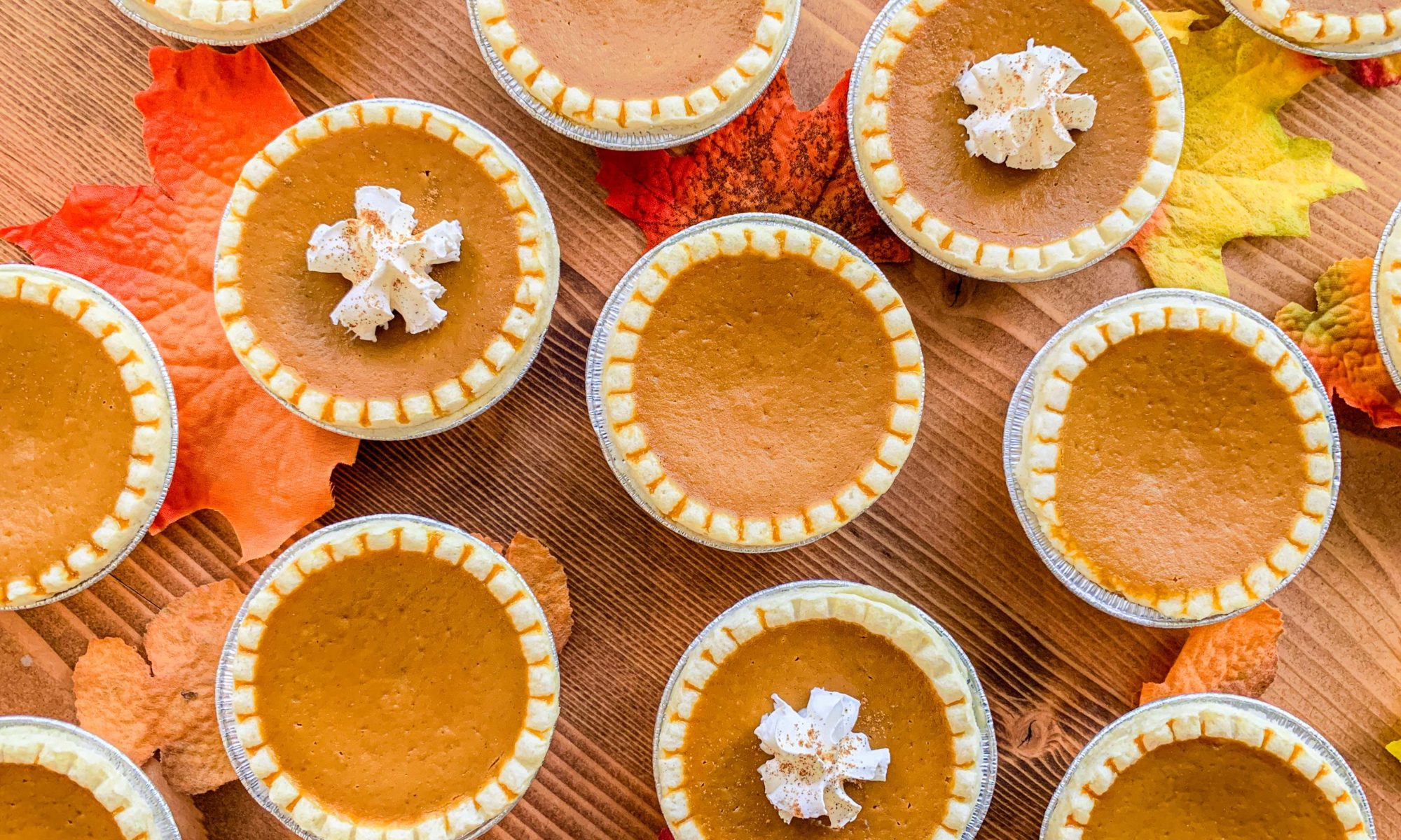 overhead shot of mini pumpkin pies, topped with whipped cream and cinnamon, on wood table top, surrounded by fall leaves