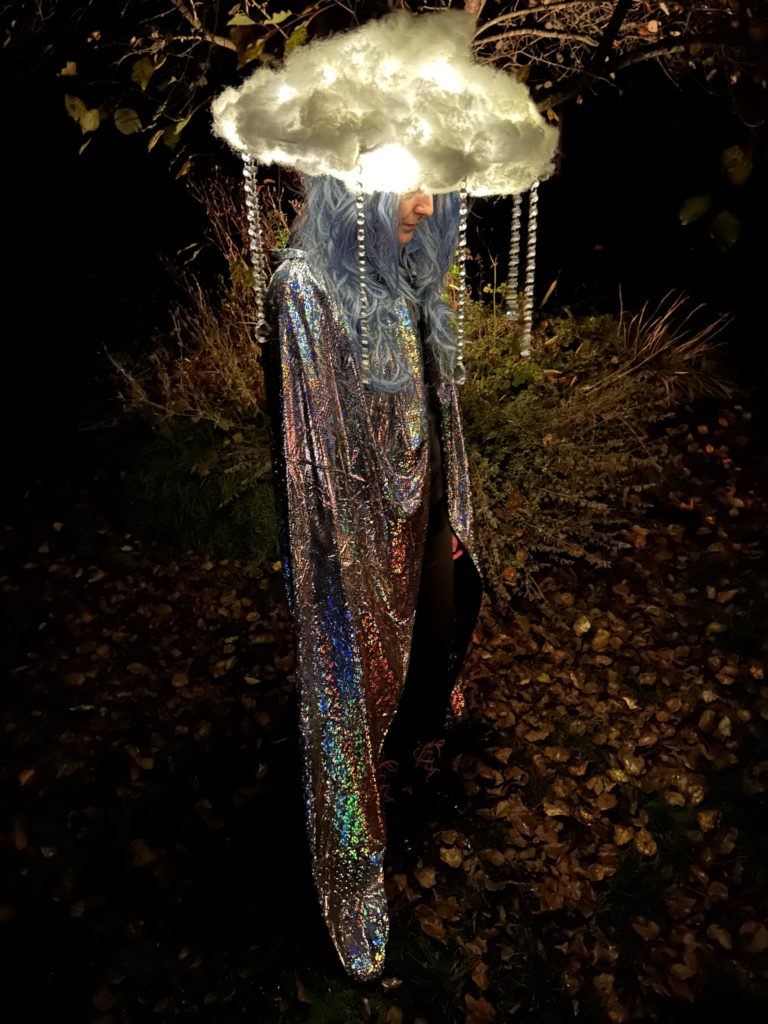 Me standing under a tree at night wearing the lit up cloud hat, a blue wig, and a silver cloak.