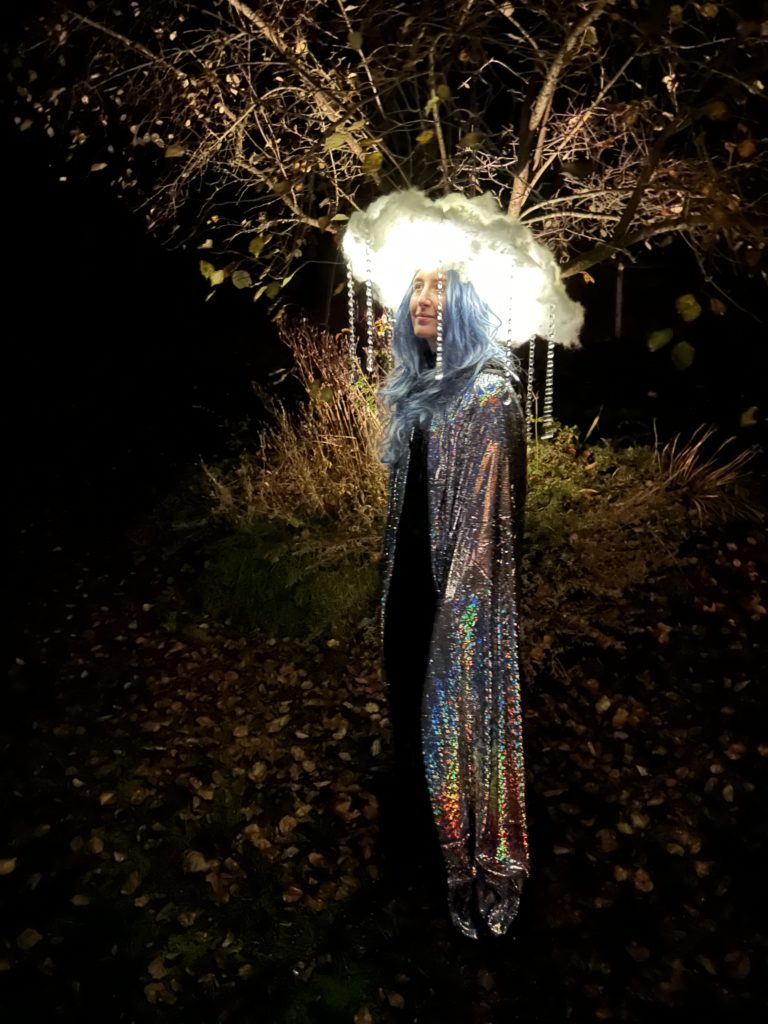 Me standing under a tree at night wearing the lit up cloud hat, a blue wig, and a silver cloak.