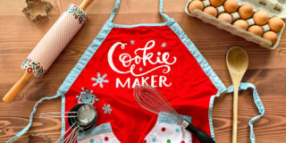 table covered in baking supplies (apron, measuring cups, whisk, rolling pin, wooden spoon, carton of eggs)